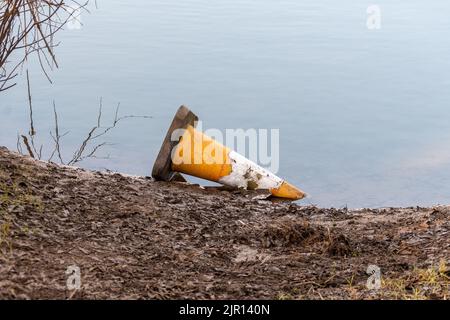 Ein schäbiger, weggeworfener Verkehrskegel, der auf seiner Seite in einem flachen Wasser neben einer schlammigen Bank liegt. Stockfoto