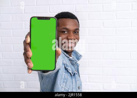 Glücklicher junger schwarzer Mann, der steht und zeigt großes Smartphone mit leerem grünen Bildschirm, der den Kopierraum für App- oder Website-Design demonstriert und darüber steht Stockfoto