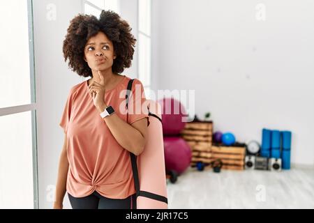 afroamerikanische Frau mit Afro-Haaren, die Yogamatte am Pilates-Raum hielt, dachte konzentriert über Zweifel mit Finger am Kinn und schaute nach oben und wunderte sich Stockfoto