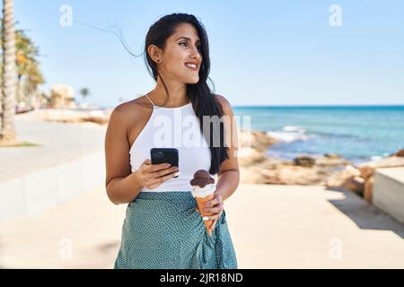 Junge hispanische Frau, die Smartphone benutzt und Eis am Meer isst Stockfoto