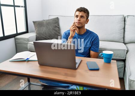 Junger gutaussehender hispanischer Mann mit Laptop auf dem Boden sitzend mit Hand auf Kinn, der über Fragen nachdenkt, nachdenklicher Ausdruck. Lächelnd mit nachdenklichem f Stockfoto