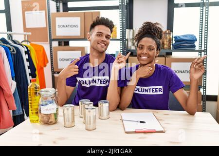 Junge interrassische Menschen, die bei Spenden ein ehrenamtliches T-Shirt tragen, stehen lächelnd und schauen auf die Kamera, die mit zwei Händen und Fingern auf den sid zeigt Stockfoto