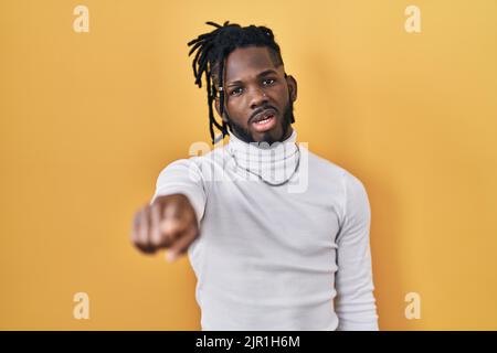 Afrikanischer Mann mit Dreadlocks, der einen Rollkragenpullover auf gelbem Hintergrund trägt und unzufrieden und frustriert auf die Kamera zeigt, wütend und wütend auf die Kamera Stockfoto