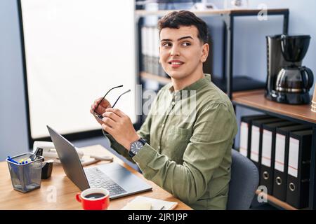 Junger nicht-binärer Mann, der im Büro eine Brille mit einem Laptop hält Stockfoto