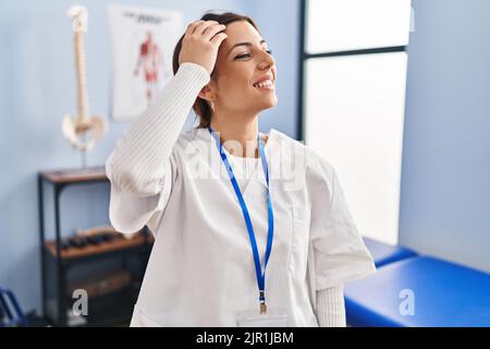 Junge Brünette Frau, die in der Klinik zur Schmerzwiederherstellung arbeitet, lächelt selbstbewusst und berührt die Haare mit der Hand nach oben, posiert attraktiv und modisch Stockfoto