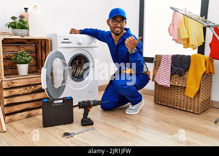 Junger indischer Techniker, der an der Waschmaschine arbeitet, lächelt mit einem glücklichen Gesicht und zeigt mit dem Daumen nach oben zur Seite. Stockfoto