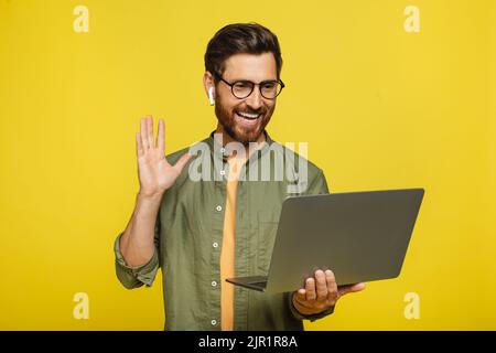 Ein glücklicher Mann mittleren Alters winkt auf den Laptop, führt einen Videoanruf über die Webcam und führt Online-Konferenzen auf gelbem Hintergrund durch Stockfoto