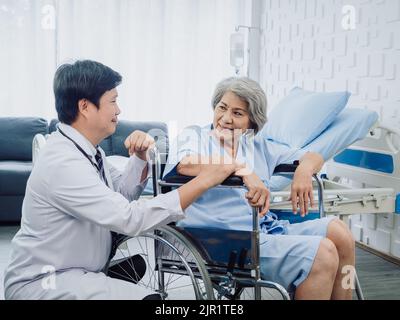 Asiatische ältere Frau Patientin in blau gekleidet, lächelnd sitzt glücklich im Rollstuhl, mit freundlicherweise Mann Arzt in weißem Anzug hält ihre Hand neben in erholen Stockfoto