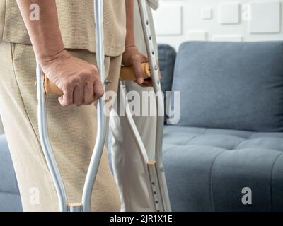 Nahaufnahme die Hände einer älteren Frau halten sich an Krücken, die zu Hause im Wohnzimmer stehen und versuchen zu gehen. Hilfe und Pflege des älteren Patienten beim Gehen mit Krücke Stockfoto