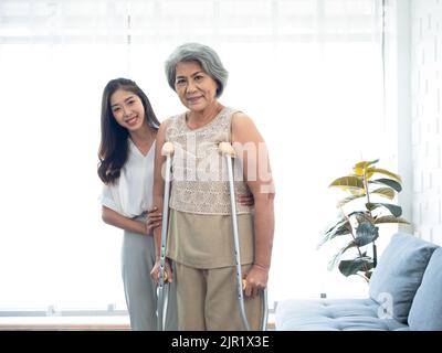 Ältere Frau, die versucht, auf Krücken zu gehen, die von jungen asiatischen Frauen in der Aufwachkammer gehalten und in den Armen unterstützt werden, um alten Frauen zu helfen und zu heilen Stockfoto