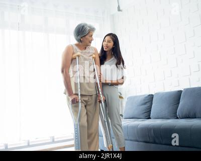 Junge lächelnde asiatische Frauen halten und unterstützen sorgfältig die Arme älterer Frauen, die versuchen, auf Krücken im Aufwachraum zu gehen und alten Frauen zu helfen, gesund zu werden Stockfoto