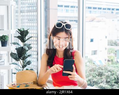 Junge schöne lächelnde asiatische Frau hält und mit Smartphone, Blick auf den Bildschirm. Glückliche asiatische Frau trägt rosa ärmellos, Sonnenbrille, s Stockfoto