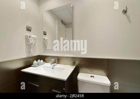 Modernes Badezimmer mit weißem Porzellanwaschbecken, dunklem Holzschrank mit rahmenlosem Spiegel an der Wand, Haartrockner mit Plug und glänzenden Fliesen Stockfoto