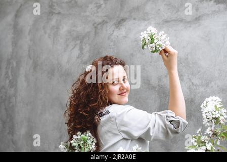 Mädchen mit langen lockigen braunen Haaren in Leinenhemd hält blühenden Ast von Apfelbaum in der Hand vor dem Hintergrund der strukturierten Betonwand. Frühjahrsblühzeit. Copy SpaceUnabhängigkeitstag der Ukraine Stockfoto