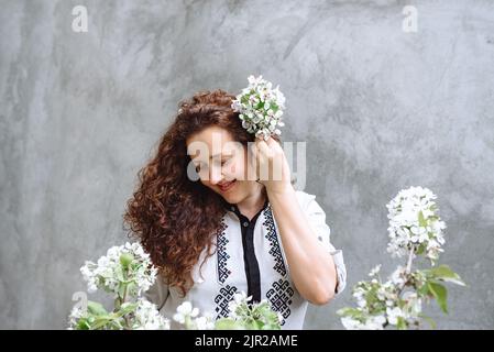 Mädchen mit lockigen Haaren in einem Hemd mit einem Downcast-Look. Frau hält blühenden Ast von Apfelbaum in der Hand vor dem Hintergrund der Betonwand. Frühjahrsblühzeit. Tag der Verfassung der Ukraine Stockfoto