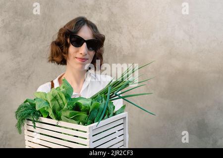 Nahaufnahme eines Mädchens, das eine weiße Holzkiste mit frischen Gemüsezwiebeln, Spinat und Dill vor dem Hintergrund einer Betonwand hält. Frau in Sonnenbrille und Hemd. Ideal für diätetischen Frühlingssalat. Speicherplatz kopieren Stockfoto