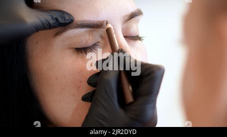 Schönheitssalon. Augenbrauenkorrektur. Nahaufnahme, Kosmetologe, Braumeister, Meister in schwarzen Handschuhen, zeichnet mit Spezialstift die Form der Augenbrauen, bevor Augenbrauenfarbstoff aufgetragen wird. Hochwertige Fotos Stockfoto