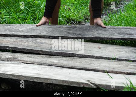 Junger lateinischer Kerl, der einige alte graue hölzerne Planken auf einem grasbewachsenen Land legt, Bau eines hölzernen Pfades oder Fußbodens. Konzept des Wohlstands. Knappe wirtschaftliche re Stockfoto