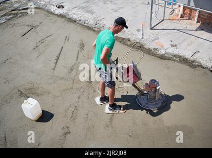 Die gesamte Länge des männlichen Arbeiters, der eine Kelle benutzte, während er im neuen Gebäude auf dem Estrichboden stielte. Mann in kurzen Hosen, der Betonoberfläche auf der Baustelle mit der Schleifmaschine für Estrich auf dem Boden fertig gestellt hat. Stockfoto