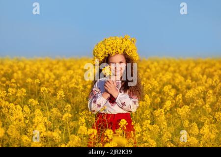 Ukrainische Mädchen in traditionellen Stickereien und gelben Kranz im Feld der gelben Blumen gegen den blauen Himmel. Betet für die Ukraine. Der Tag der Unabhängigkeitsfahne der Ukraine. Tag der Verfassung. Symbole der Ukraine. Stockfoto