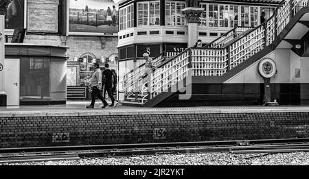 Die Passagiere gehen auf den Stufen zu einer Fußgängerbrücke des Bahnhofs. Auf der Plattform befindet sich eine Wiegemaschine und darüber ein Café. Stockfoto