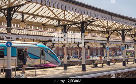 Ein Zug ruht unter einem alten historischen Vordach aus dem 19.. Jahrhundert. Passagiere sind auf dem Bahnsteig und ein Jugendlicher fotografiert die Lokomotive. Stockfoto