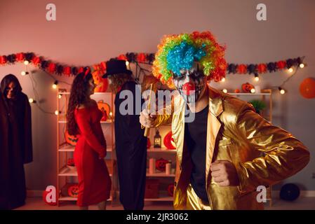Junger Mann, gekleidet als gruseliger böser Clown mit Axt auf Halloween-Party mit Freunden Stockfoto
