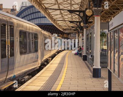 Ein Zug steht neben einem Bahnhofssteig. Ein historisches Baldachin aus dem 19.. Jahrhundert, das von Säulen gestützt wird, ist darüber. Ein Passagier sitzt auf einer Bank und einem Adve Stockfoto