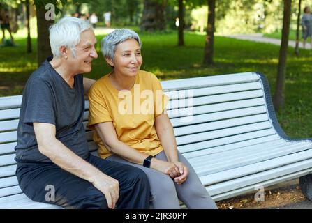 Ziemlich multirassische ältere Menschen 60s Paar sitzen auf der Bank im Park nach einem Spaziergang, Morgenspaziergang lächelnd Genießen Sie Gespräche entspannt im Freien an sonnigen Sommertag Stockfoto