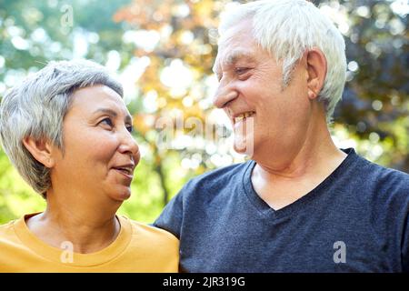 Optimistische attraktive multirassische ältere Menschen 60s sportliche Paare posieren im Park lächelnd und blicken sich mit Zärtlichkeit und Liebe an. Happy Endless Marriag Stockfoto