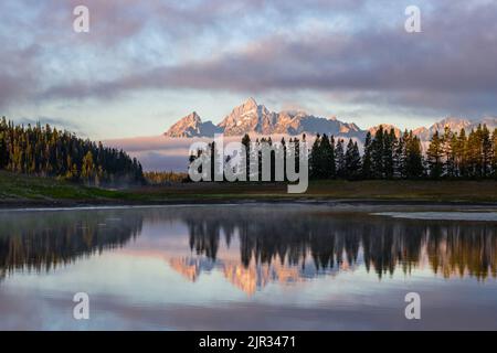 Morning Mists teilen sich, um die Teton Range in all ihrer Pracht zu enthüllen, die sich in stillem Wasser widerspiegelt Stockfoto