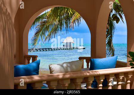 Tropischer Blick auf einen Palapa am Ende eines Piers über dem Wasser, von einem Pavillon auf einem Strand auf Ambergris Caye, San Pedro, Belize aus gesehen. Stockfoto