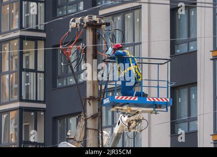 Ein Elektriker, der in der Wiege eines Turmlifts steht, montiert Drähte auf einem Mast vor dem Hintergrund eines Hochhauses Stockfoto