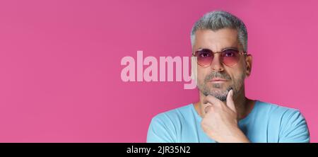 Nachdenklicher, hübscher Mann 30s 40s mit pinker Sonnenbrille und blauem T-Shirt, der sein Kinn mit einem nachdenklichen, konzentrierten Blick auf rosa Hintergrund berührt. Grauhaarige reifen Mann Studio geschossen. Stockfoto