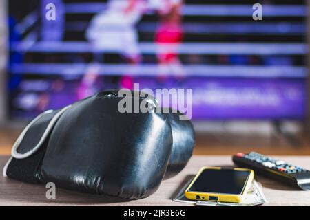 Schwarze Boxhandschuhe, ein Handy mit einem gelben Gehäuse über Dollarscheinen und eine TV-Fernbedienung auf einem Holztisch. Im Hintergrund, außer Fokus, ein b Stockfoto