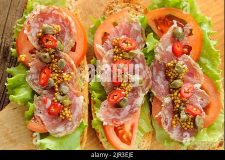 Sandwiches mit Schinken, Salatblättern, Chili, Tomaten, Kapern, französischer Senf auf Holzhintergrund horizontal Stockfoto