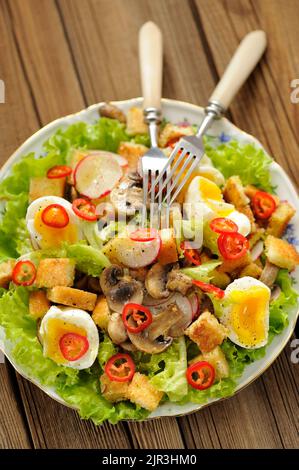 Salat Caesar mit Pilzen, Eiern, Chili und Rettich mit zwei Gabeln auf Holzhintergrund vertikal Stockfoto