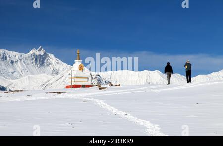 Blick auf die weiße buddhistische Stupa in der Nähe des Eissees (Kicho Tal) und der Annapurna-Reihe mit zwei Touristen, den Berg Gangapurna oder Ganggapurna um die Annapurna-Rennstrecke Stockfoto