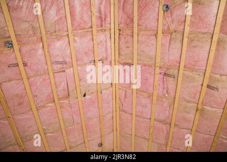 Unvollendete Decke mit Holzlatten und freiliegender rosafarbener Glasfaserisolierung in Wohnhäusern. Stockfoto