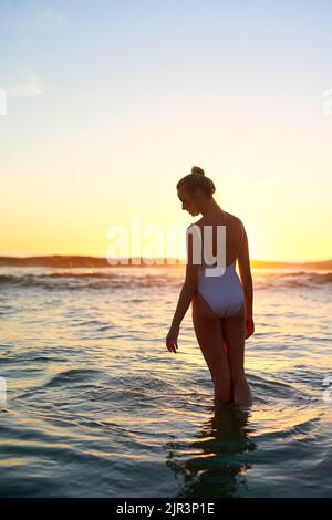 Der Strand lässt mich all die guten Erinnerungen wieder aufleben: Eine junge Frau, die bei Sonnenuntergang im Wasser steht. Stockfoto