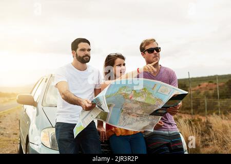 Roadtrip-Zeit. Eine Gruppe junger Freunde, die eine Straßenkarte halten und planen, wohin sie gehen sollen, während sie neben einer Straße stehen. Stockfoto