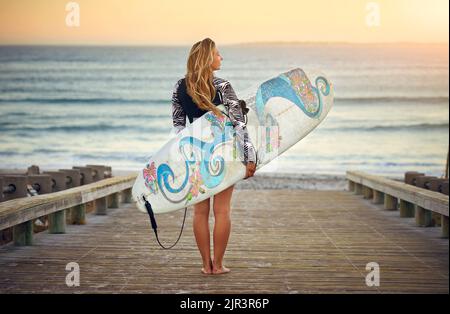 Ozeangedanken. Rückansicht einer attraktiven jungen Surferin, die mit ihrem Surfbrett über den Ozean blickt. Stockfoto