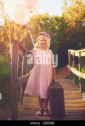 Es ist Zeit, die Welt rosa zu malen. Ein glückliches kleines Mädchen hält Ballons und einen Koffer, während es mitten auf einer Brücke steht. Stockfoto