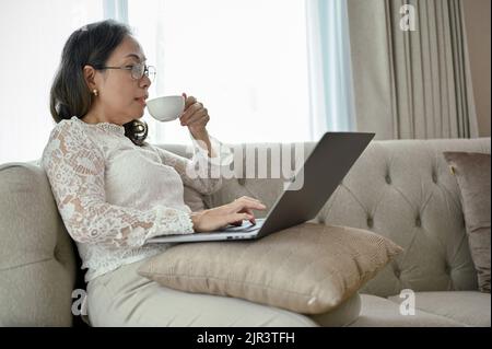Eine erfolgreiche asiatische Geschäftsfrau mittleren Alters oder weibliche Chefin schlürft heißen Tee oder Kaffee, während sie mit einem Laptop ihre Geschäftsaufgaben in verwaltet Stockfoto