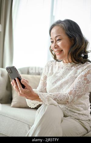 Portrait, eine attraktive und wunderschöne asiatische Frau mittleren Alters nutzt ihr Smartphone, während sie sich im Wohnzimmer entspannt. Stockfoto