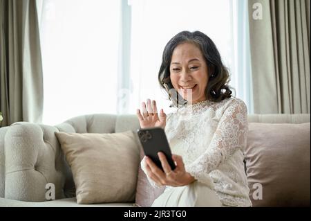Fröhliche und fröhliche Frau im asiatischen Alter genießen es, bei einem Videoanruf mit ihrem Enkelkind zu sprechen, entspannt sich im Wohnzimmer sitzend. Stockfoto