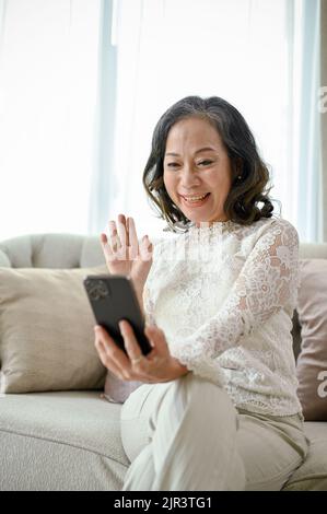 Portrait, Happy Asian Middle-aged woman nutzt ihr Smartphone für Videoanruf, winkt ihre Hand, um ihre Freundin zu grüßen und zu grüßen. Stockfoto