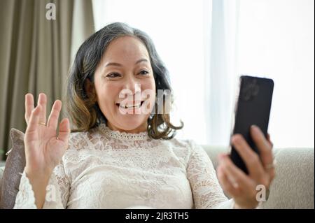 Die wunderschöne und glückliche asiatische Frau mittleren Alters nutzt ihr Smartphone, um mit ihren Freunden im Wohnzimmer zu telefonieren. Stockfoto