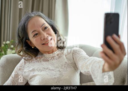 Wunderschöne und glückliche asiatische Frau mittleren Alters verwendet ihr Smartphone, um ein Selfie in ihrem minimalen Wohnzimmer zu machen. Stockfoto