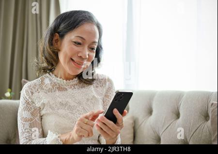 Eine fröhliche und lächelnde Asiatin verwendet ihr Smartphone, um mit ihren Freunden zu schreiben, während sie sich auf dem Sofa im Wohnzimmer entspannt. Stockfoto